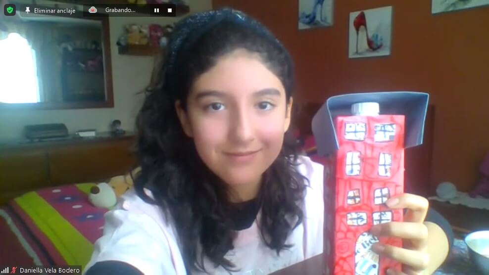 Captura de clase en vivo: niña mostrando su edificio hecho con caja de cartón reciclada y pintada de rojo