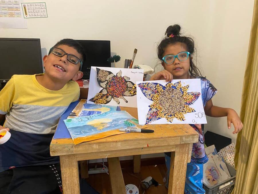 Dos niños junto a sus obras de arte con semillas y granos: una mariposa y una flor hechas con arroz y frijoles