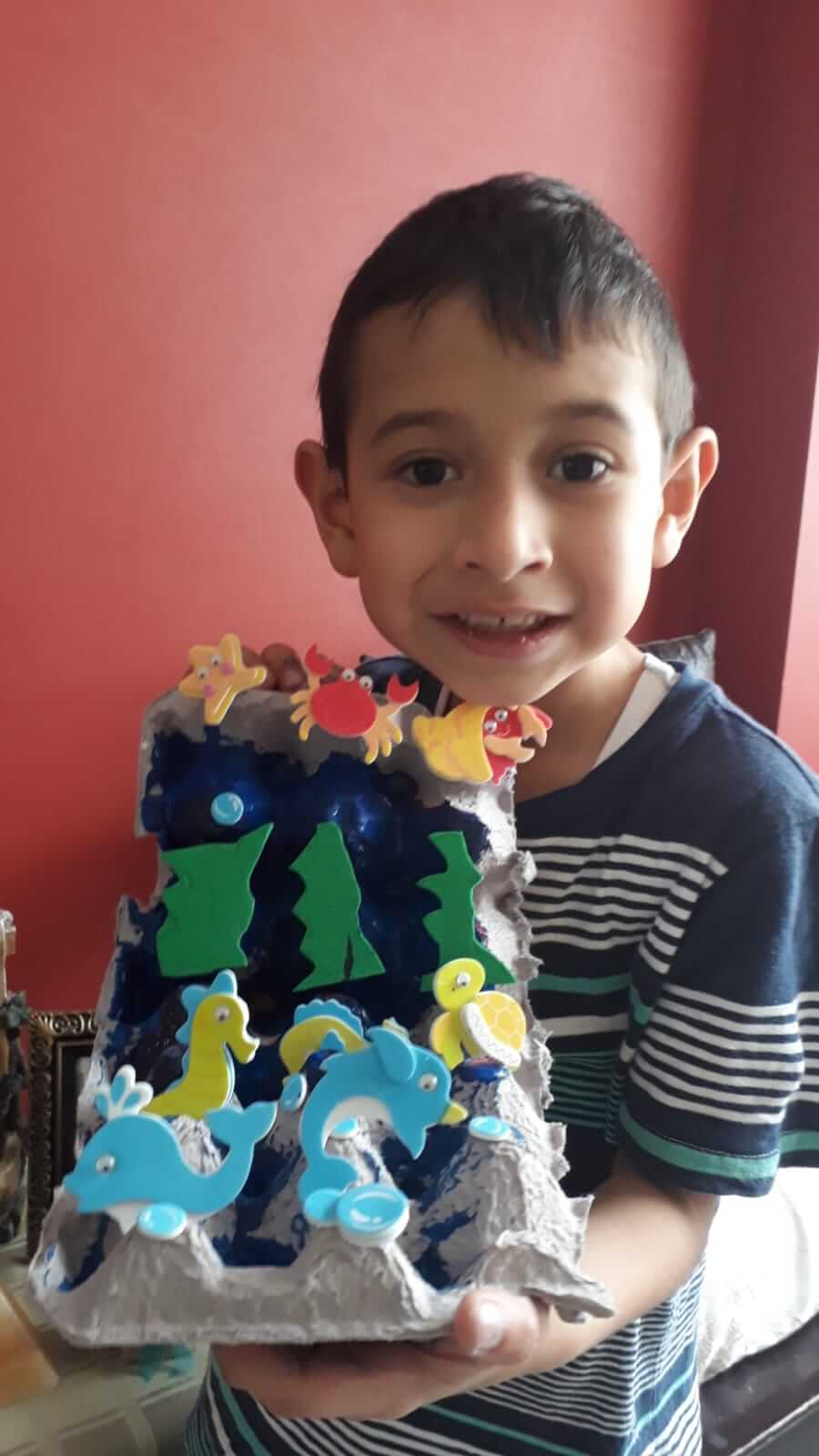 Niño sonriente mostrando su diorama del océano hecho con cartón de huevos, peces, delfín, cangrejo y tortuga