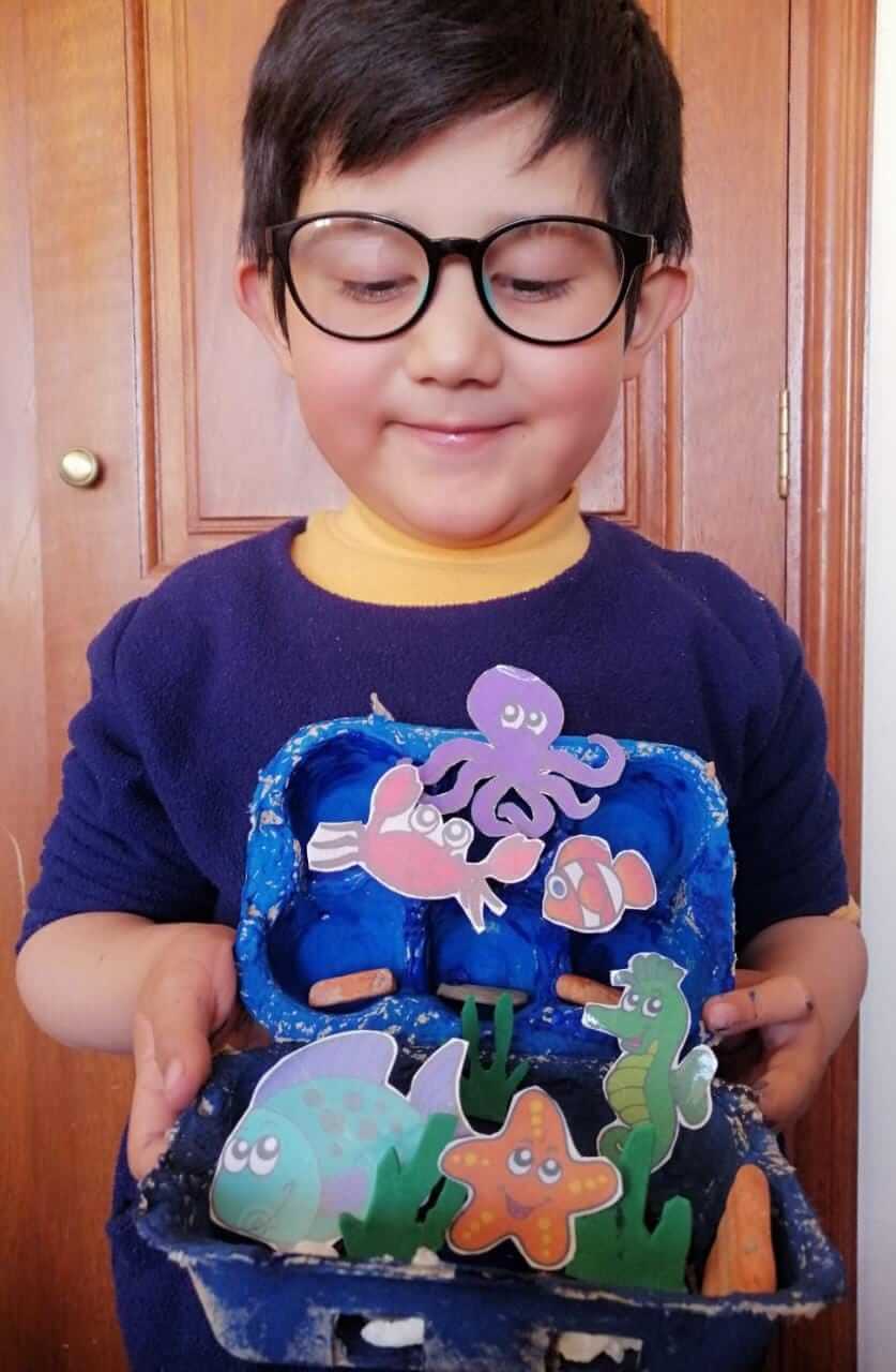 Niño con lentes sosteniendo su acuario hecho con cartón de huevos azul, con pulpo, pez y caballito de mar