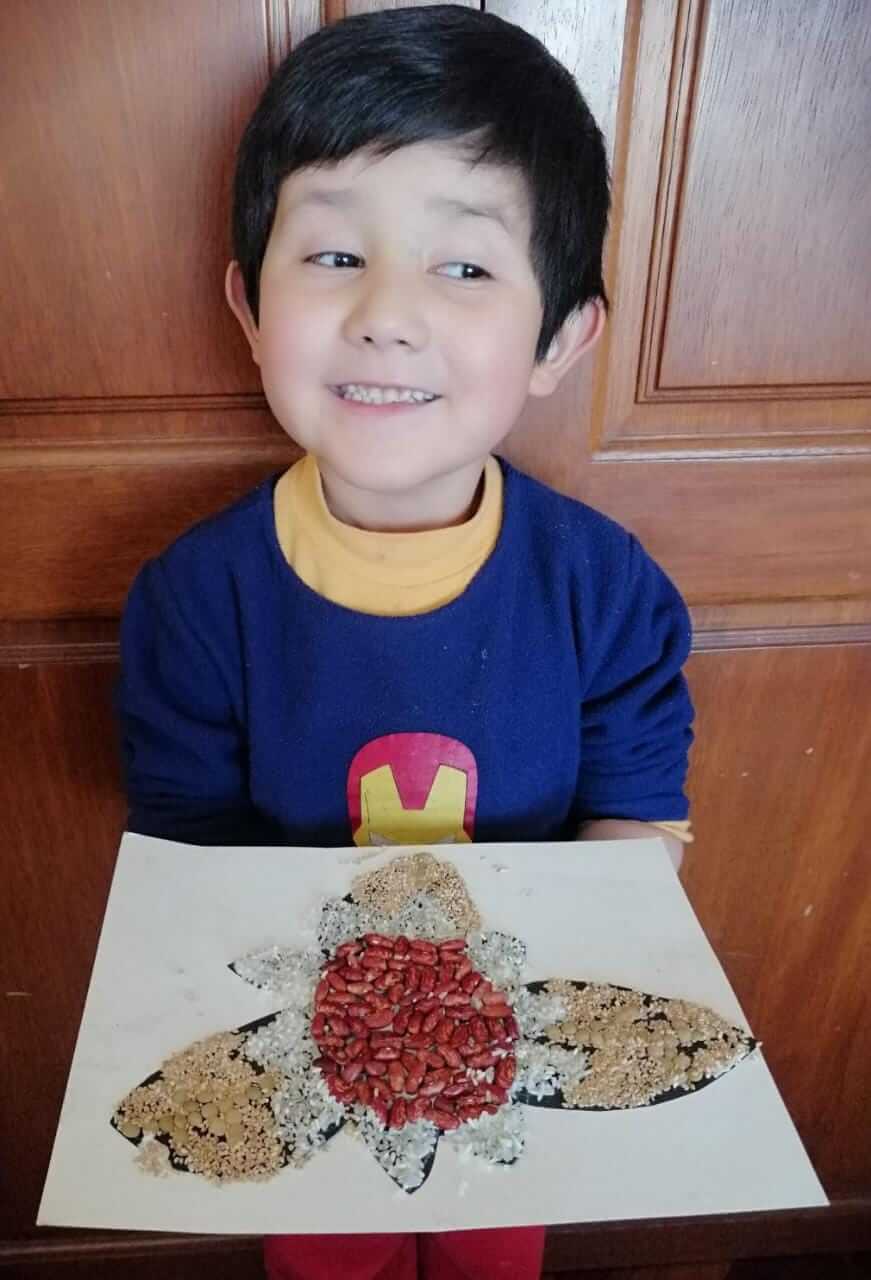 Niño mostrando su obra de arte con semillas y granos (arroz, frijoles) formando una flor en papel