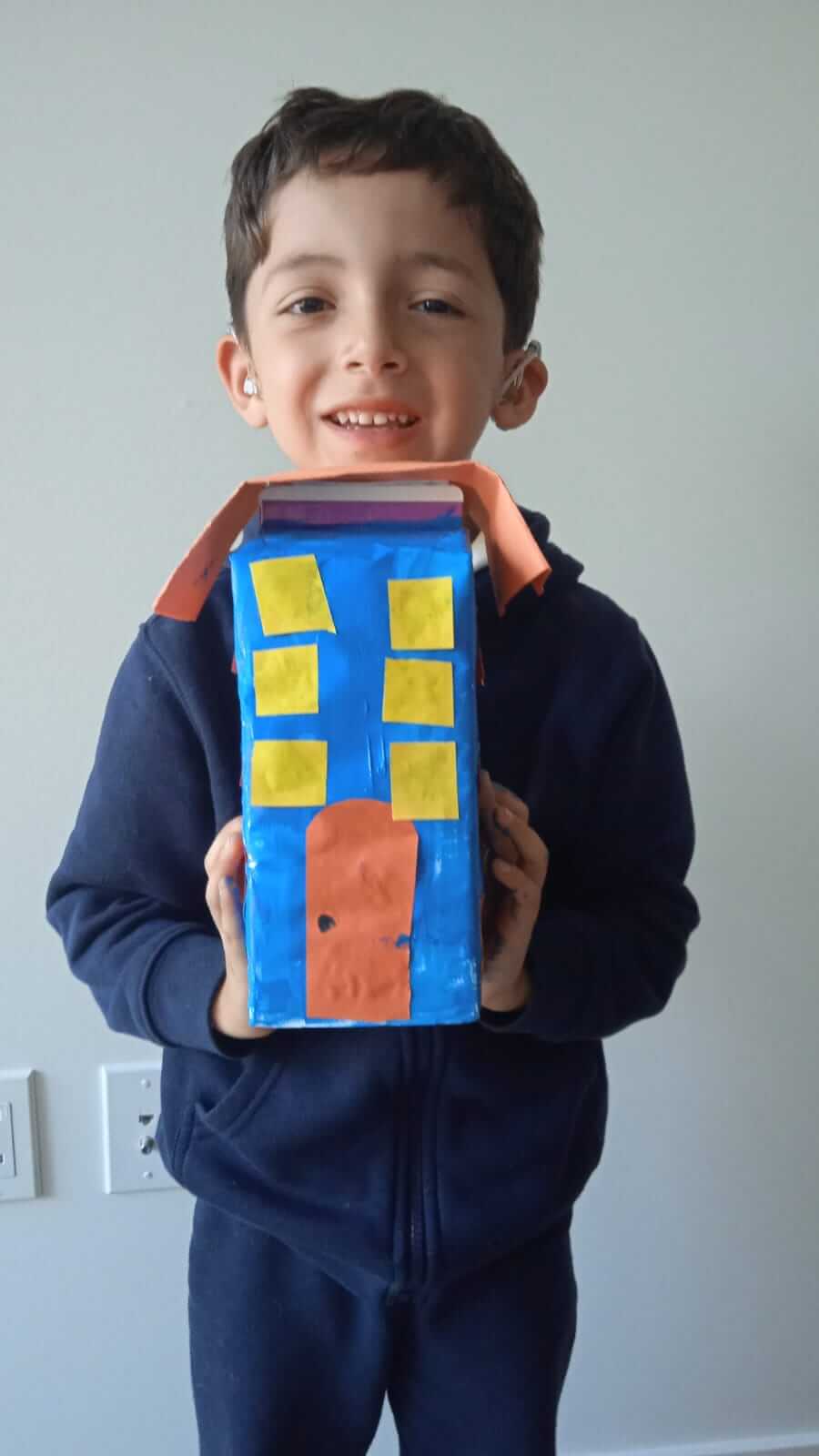 Niño sonriente mostrando su casa hecha con caja de cartón pintada de azul, con ventanas amarillas y puerta naranja