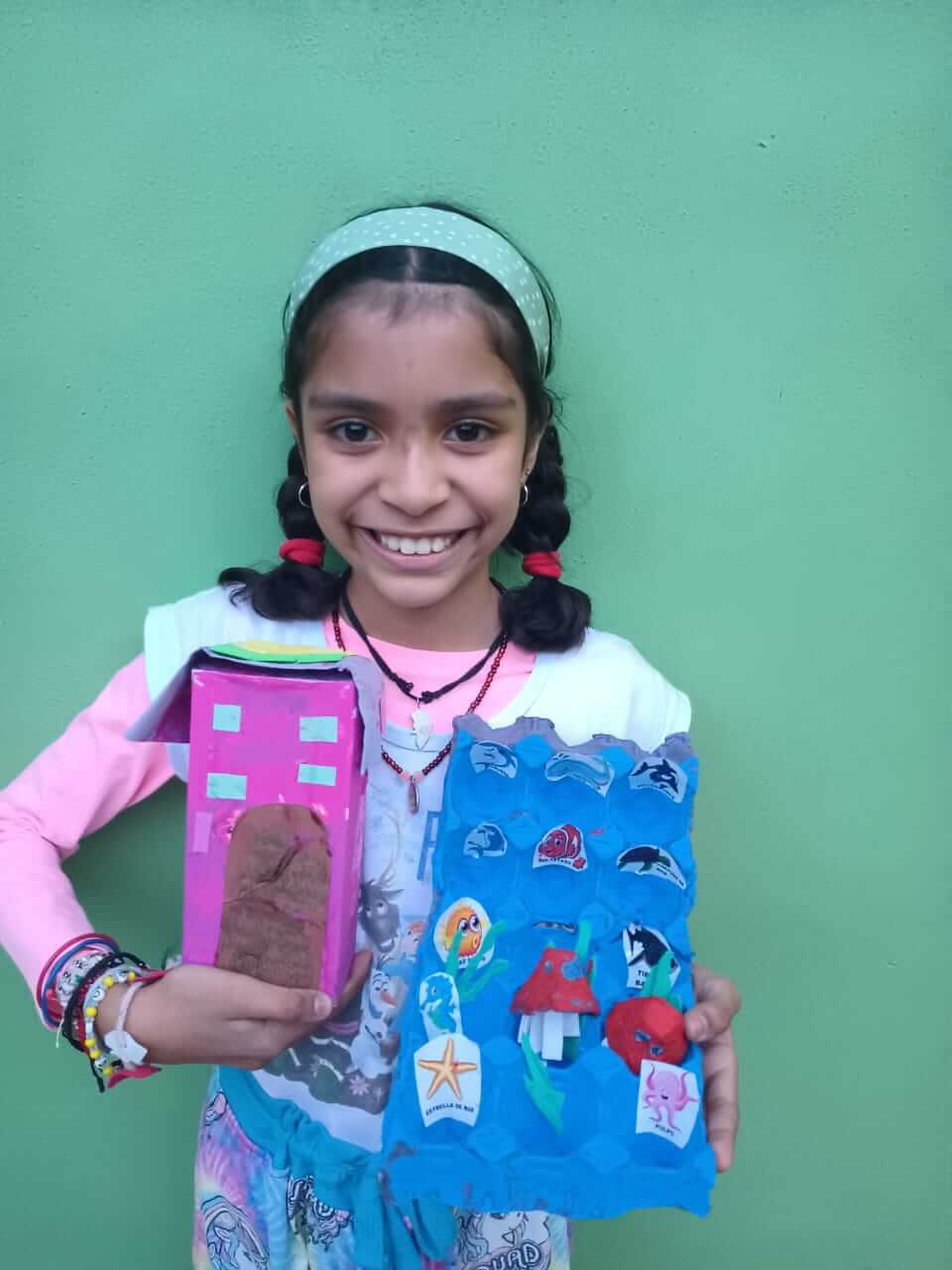 Niña mostrando dos manualidades: una casita de cartón rosa y un acuario con cartón de huevos y figuras marinas